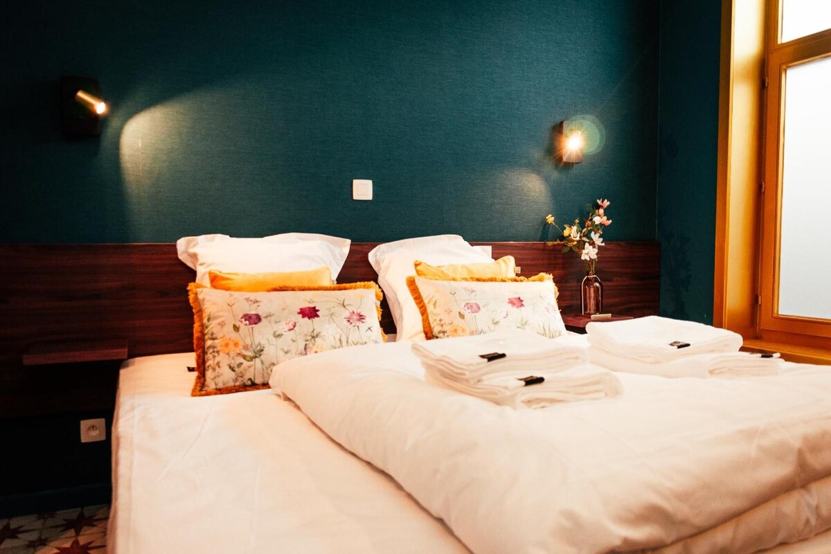 Una cama bien hecha con almohadas florales y toallas dobladas, una mesita de noche con un florero y una lámpara de pared.