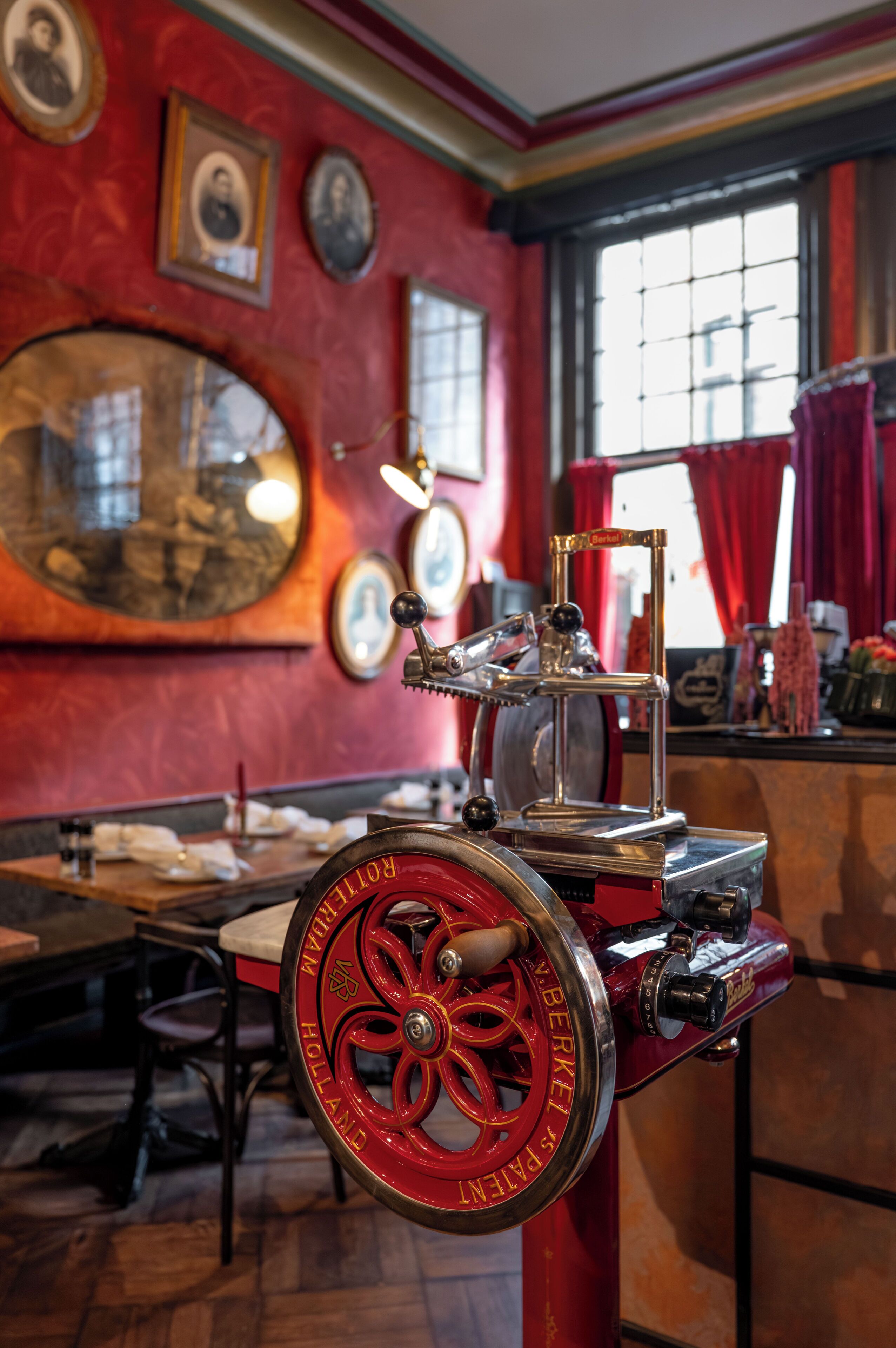 A vintage espresso machine in a room with red walls, framed pictures, and a window with red curtains.