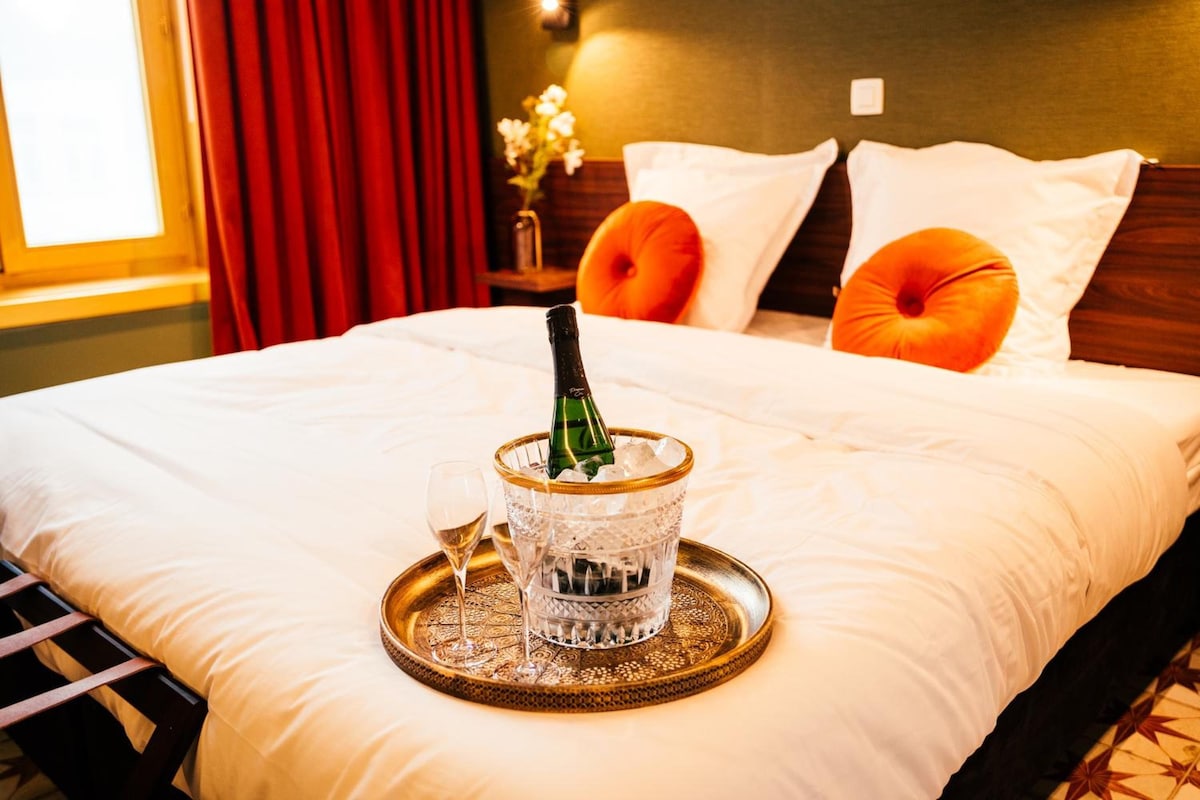 Ein ordentlich bezogenes Bett mit Champagnerflasche und Gläsern auf einem Tablett, ein Nachttisch mit Blumen in einer Vase und ein Fenster mit Vorhängen.