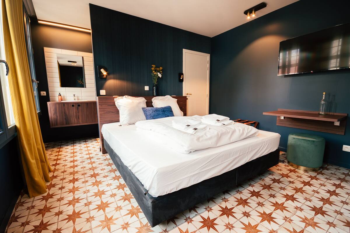 A hotel room with a large bed, a wooden dresser, a mirror, a TV, and a small green stool.