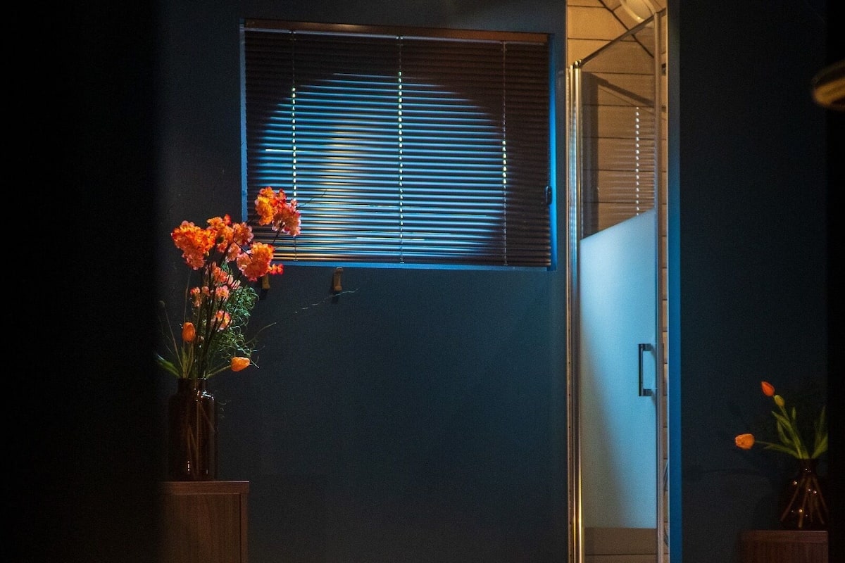 A bedroom with a bed, a window with blinds, a vase with flowers, and a patterned floor.