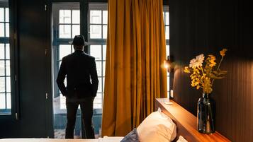 A hotel room with a bed, a nightstand, a vase with flowers, and a window with curtains.