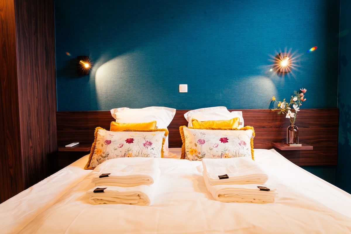 A neatly made bed with floral pillows and folded towels, a bedside table with a vase of flowers, and a nightstand with a remote control.