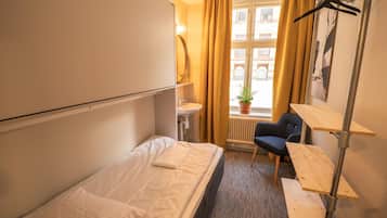A small hotel room with a single bed, a desk, a chair, a radiator, a window with curtains, and a hanging coat rack.