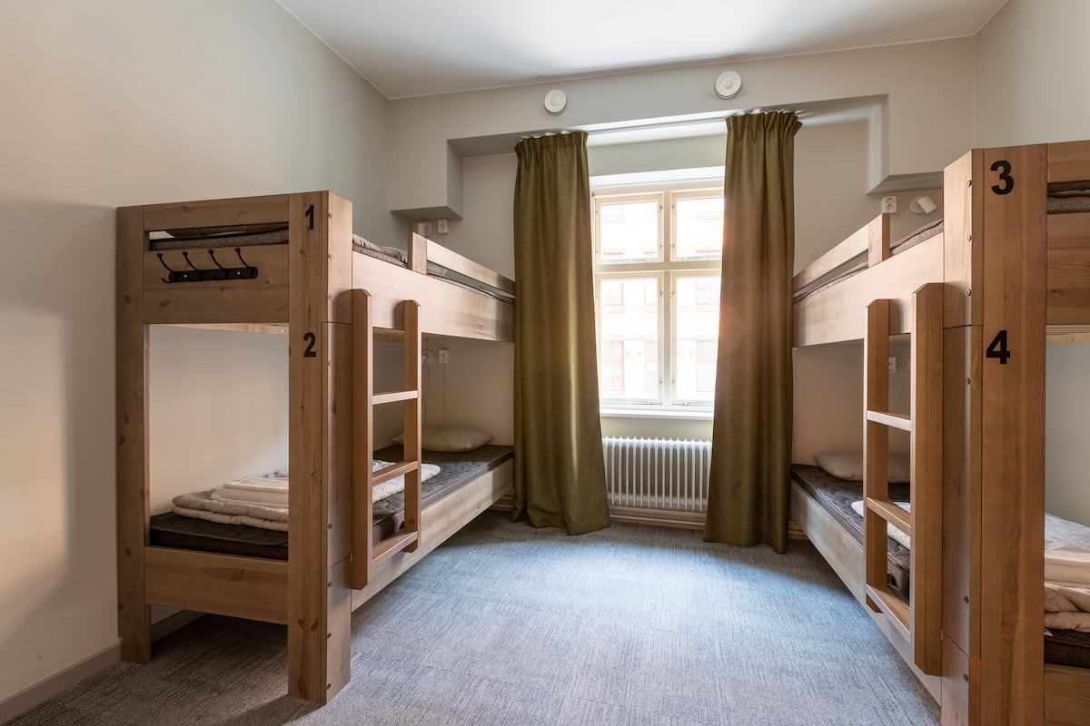 A room with four bunk beds, numbered 1 to 4, wooden walls, a window with curtains, and a radiator.