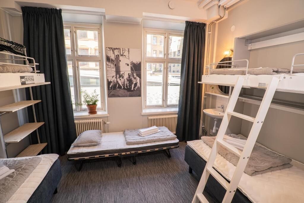 A dormitory room with bunk beds, a window with curtains, a radiator, and a poster on the wall.