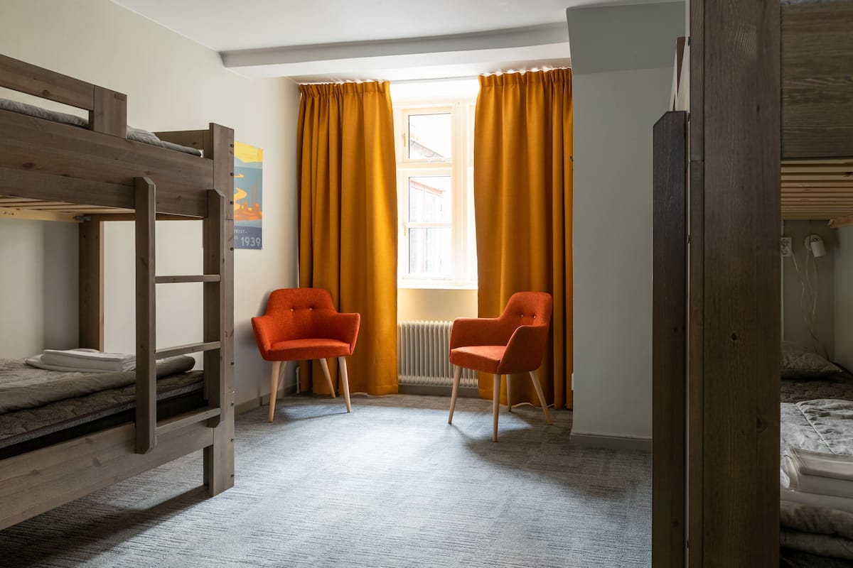 A room with bunk beds, two orange chairs, a window with yellow curtains, and a radiator.