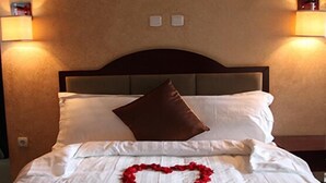 Cama bem arrumada com lençóis brancos e travesseiro marrom, com um enfeite em formato de coração sobre a cama, e luminárias fixadas na parede de cada lado.