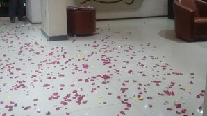 Uma área de recepção com piso ladrilhado coberto de pétalas de rosa, um balcão de atendimento e uma parede com relógios e a placa 'Reception'.