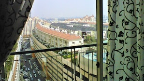 Blick aus einem Fenster mit gemusterten Vorhängen auf einen Stadtblick mit Wohngebäuden, einer Straße mit parkenden Autos und einem großen Gebäude mit einer gewölbten Dachform.
