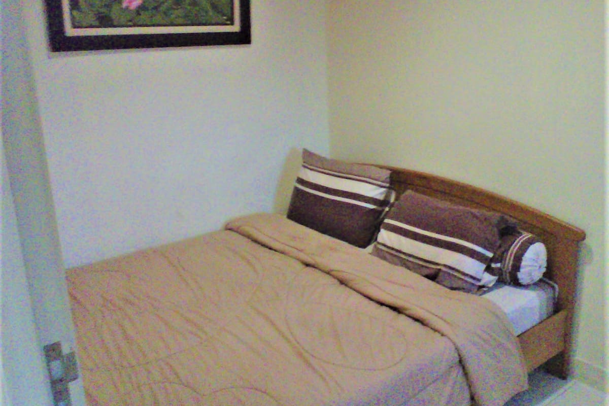 A small, neatly made bed with a beige blanket and two striped pillows, a framed picture on the wall, and a plain white wall.