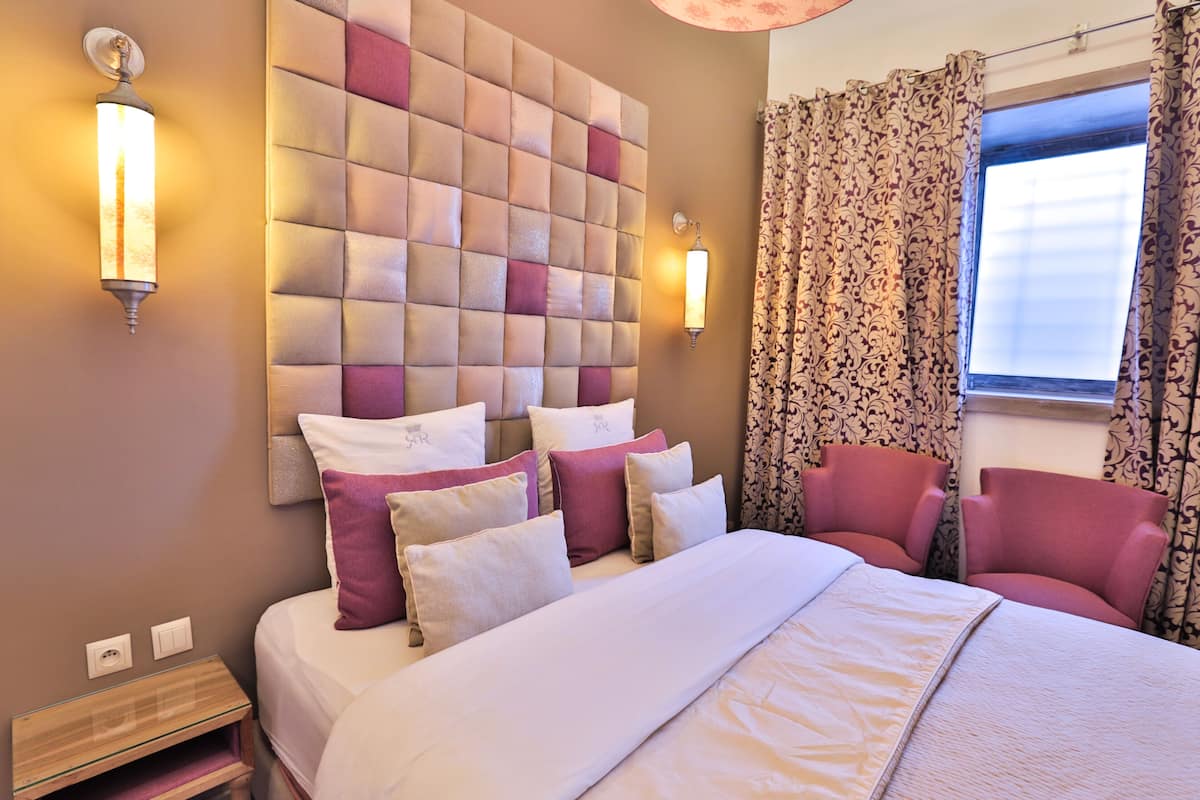 Habitación de hotel con cabecera capitonada, varias almohadas, cama, mesita de noche, silla y ventana con cortinas.