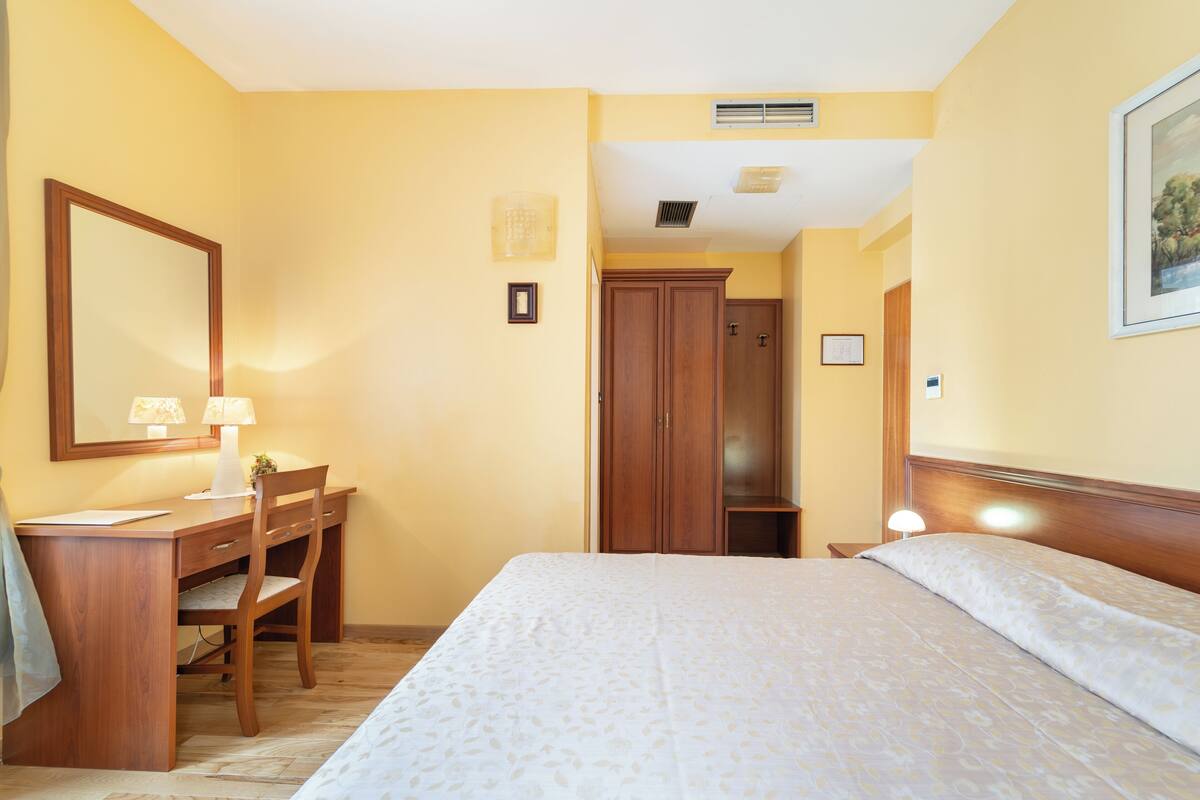 A hotel room with a bed, a desk with a chair, a mirror, and a door leading to another room.
