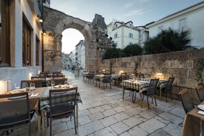 An outdoor dining area with stone tables and chairs, an archway, and buildings in the background.