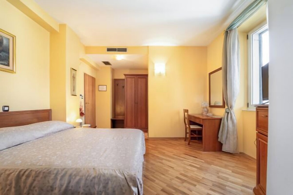 Habitación de hotel con cama, escritorio, silla, armario y ventana con cortinas.