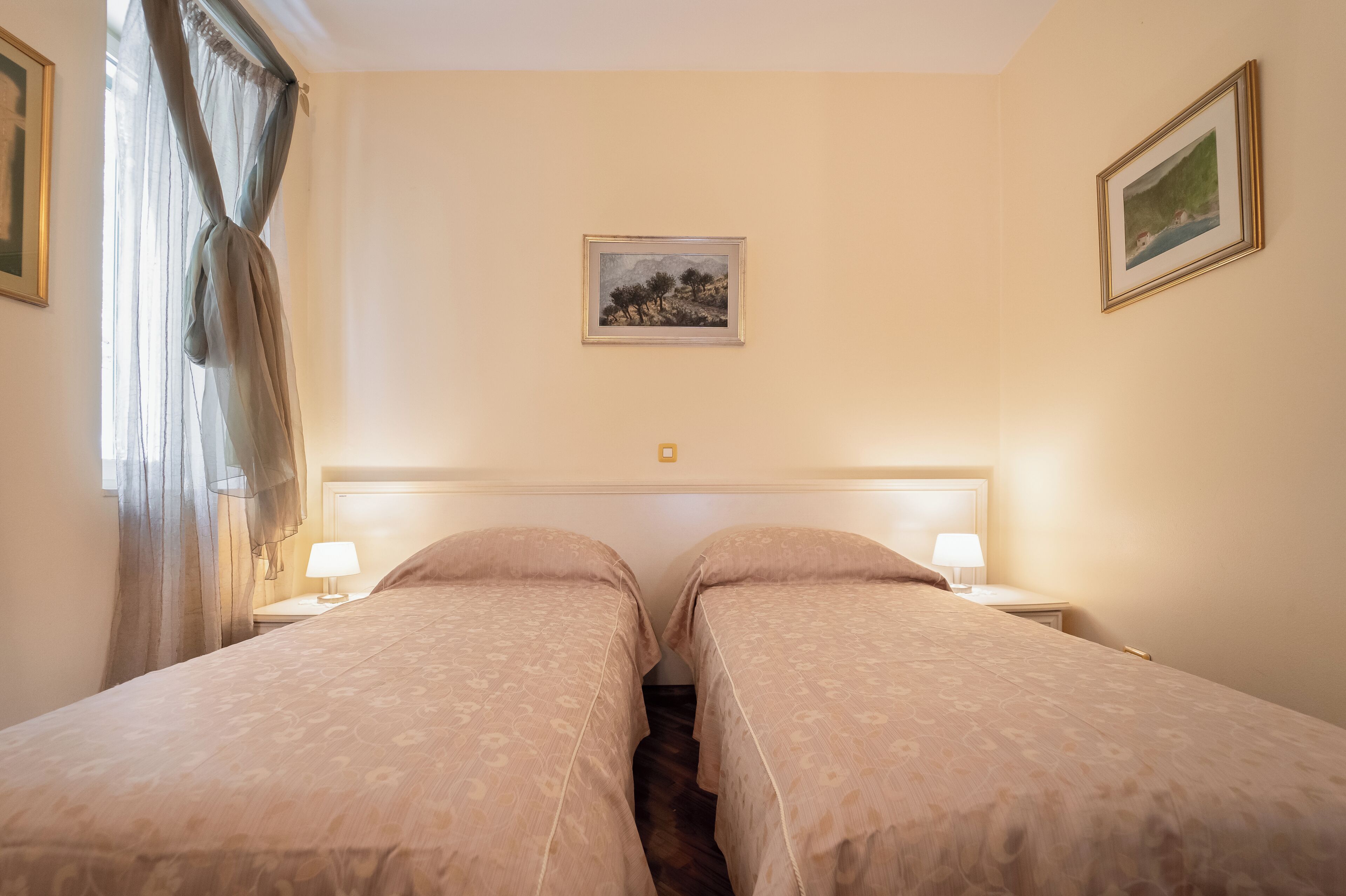 Une chambre d’hôtel avec deux lits, une tête de lit, une table de chevet avec une lampe, une fenêtre avec des rideaux légers et un tableau encadré au mur.