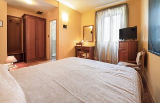 A hotel room with a large bed, a desk with a chair, a wardrobe, a television on a dresser, and a window with curtains.