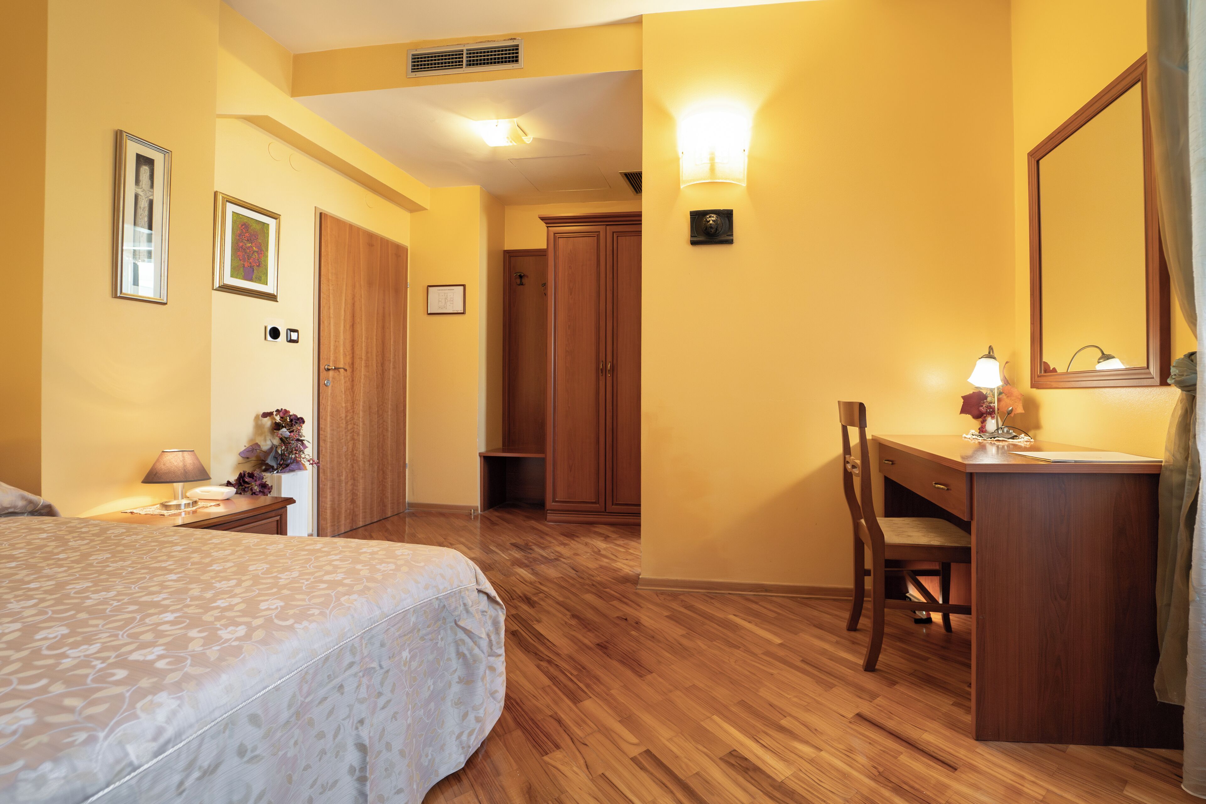 A hotel room with a bed, a desk, a chair, a mirror, and a wall-mounted light fixture.