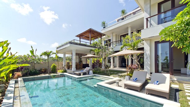 A modern pool area with lounge chairs and umbrellas, surrounded by lush greenery and a multi-level building with balconies. - Casa de Balangan by Exotiq Villa Holidays (Jimbaran)