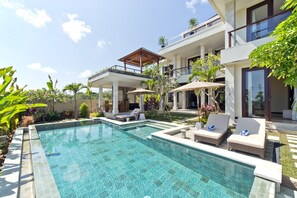 A modern pool area with lounge chairs and umbrellas, surrounded by lush greenery and a multi-level building with balconies. - Casa de Balangan by Exotiq Villa Holidays (Jimbaran)