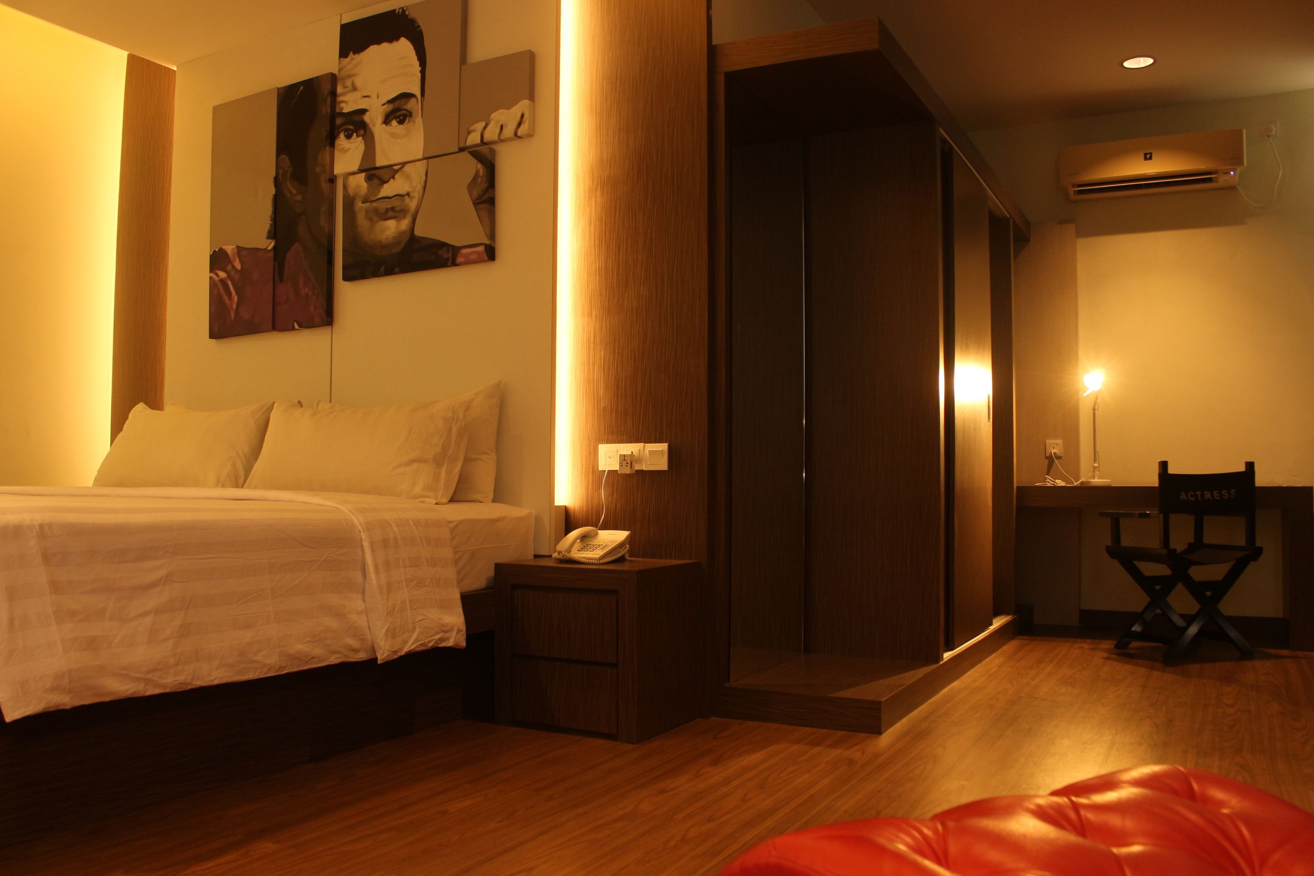 A hotel room with a bed, bedside table, lamp, wardrobe, and a red chair.