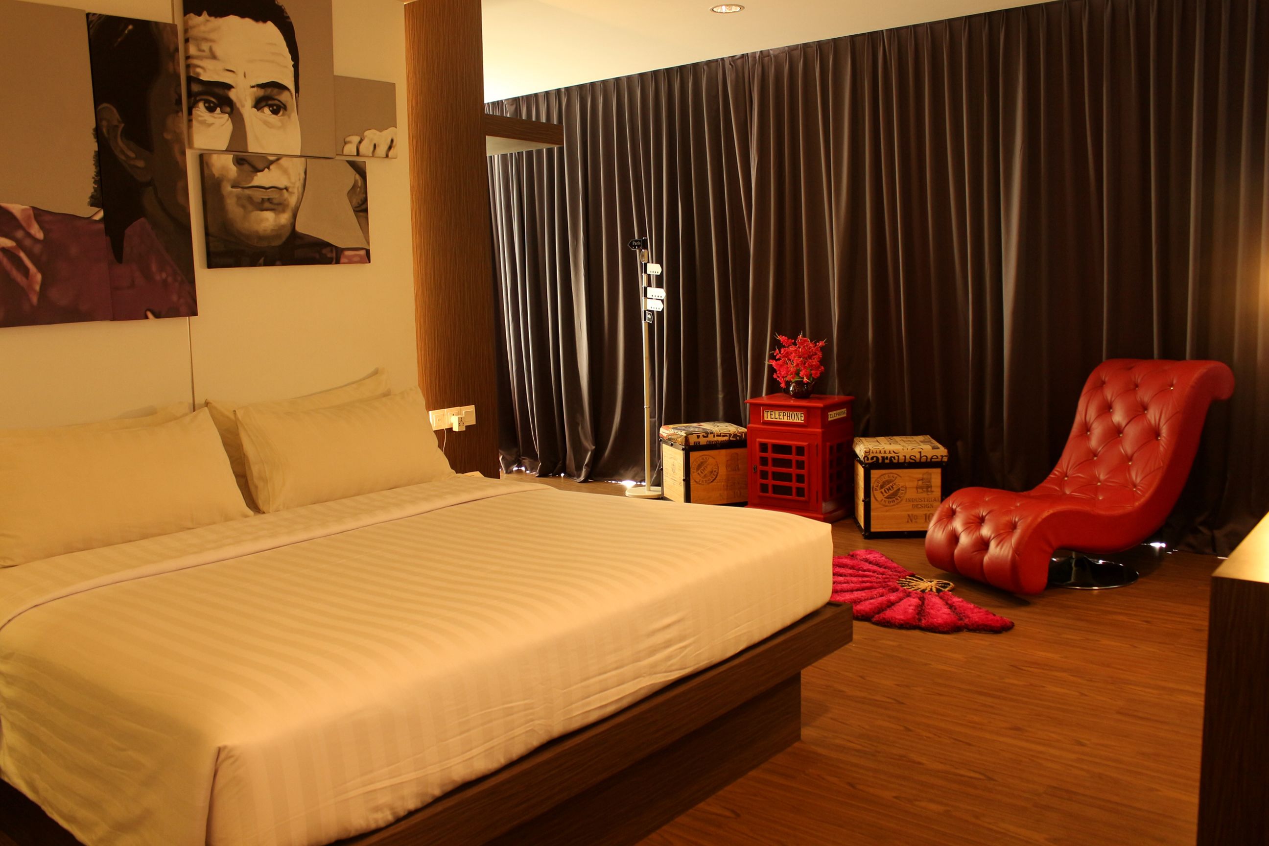 Habitación de hotel con una cama grande, un sillón rojo, una mesita de noche con un florero y un ventanal con cortinas.
