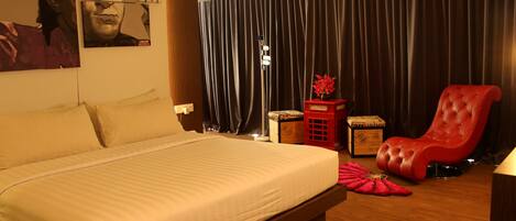 Habitación de hotel con una cama grande, un sillón rojo, una mesita de noche con un florero y un ventanal con cortinas.