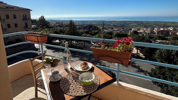 A balcony with a table set for two, potted plants, and a view of the cityscape. - B&B La finestra sulla valle (Agrigento)