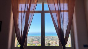 A window with sheer curtains, offering a view of a coastal landscape with buildings and greenery. - B&B La finestra sulla valle (Agrigento)
