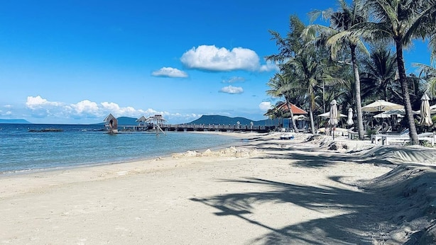 A beach with a pier, palm trees, and beach chairs. - Gold Coast Phu Quoc Resort (Phu Quoc)