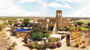 A large, historic building with a prominent tower, surrounded by a landscaped garden with a swimming pool. - Mihir Garh (Rohat)