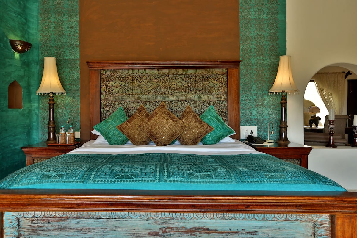 A bed with a patterned bedspread, a wooden headboard, and decorative pillows. There are two bedside tables with lamps and a wall tapestry.