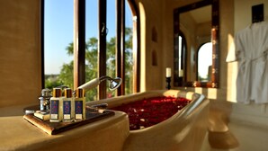 A bathtub filled with rose petals, a set of toiletries, and a window with a view of palm trees. - Mihir Garh (Rohat)