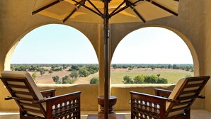A patio with two wooden chairs and a table, a central umbrella, and a view through two arches. - Mihir Garh (Rohat)
