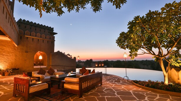 An outdoor seating area with stone flooring, a wooden bench, and a table with cushions. - Mihir Garh (Rohat)