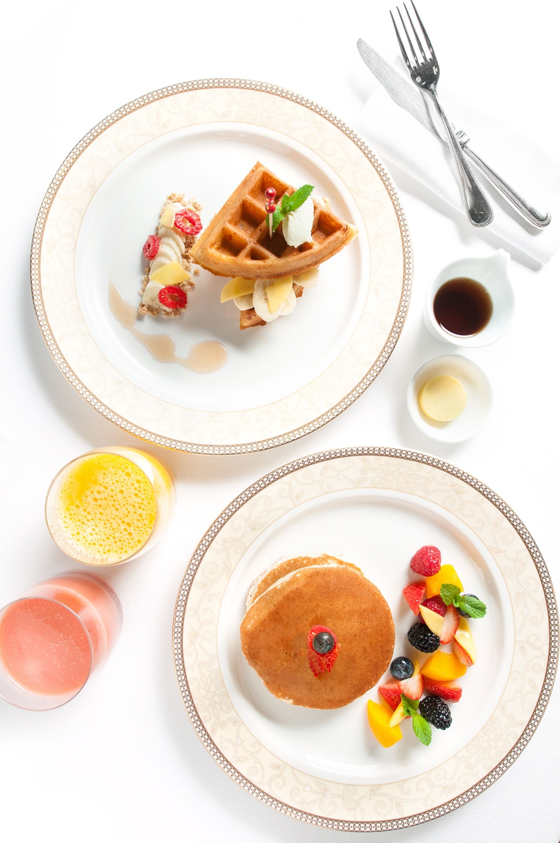 A breakfast setting with waffles, pancakes, and fruit on plates, accompanied by syrup and a glass of juice.