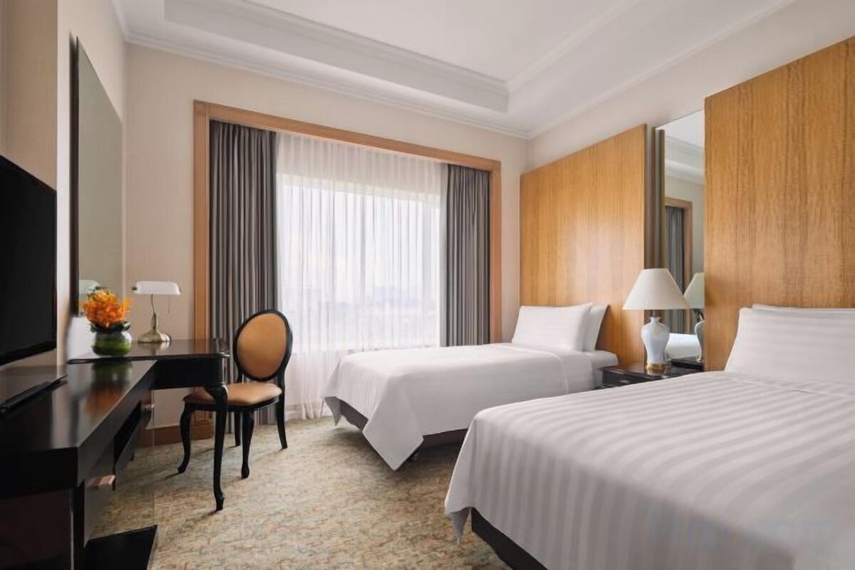 A hotel room with two beds, a desk, a chair, a TV, and a window with curtains.