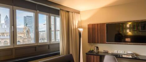 A hotel room with a large window offering a city view, a bed, a desk with a chair, and a flat-screen TV mounted on the wall.