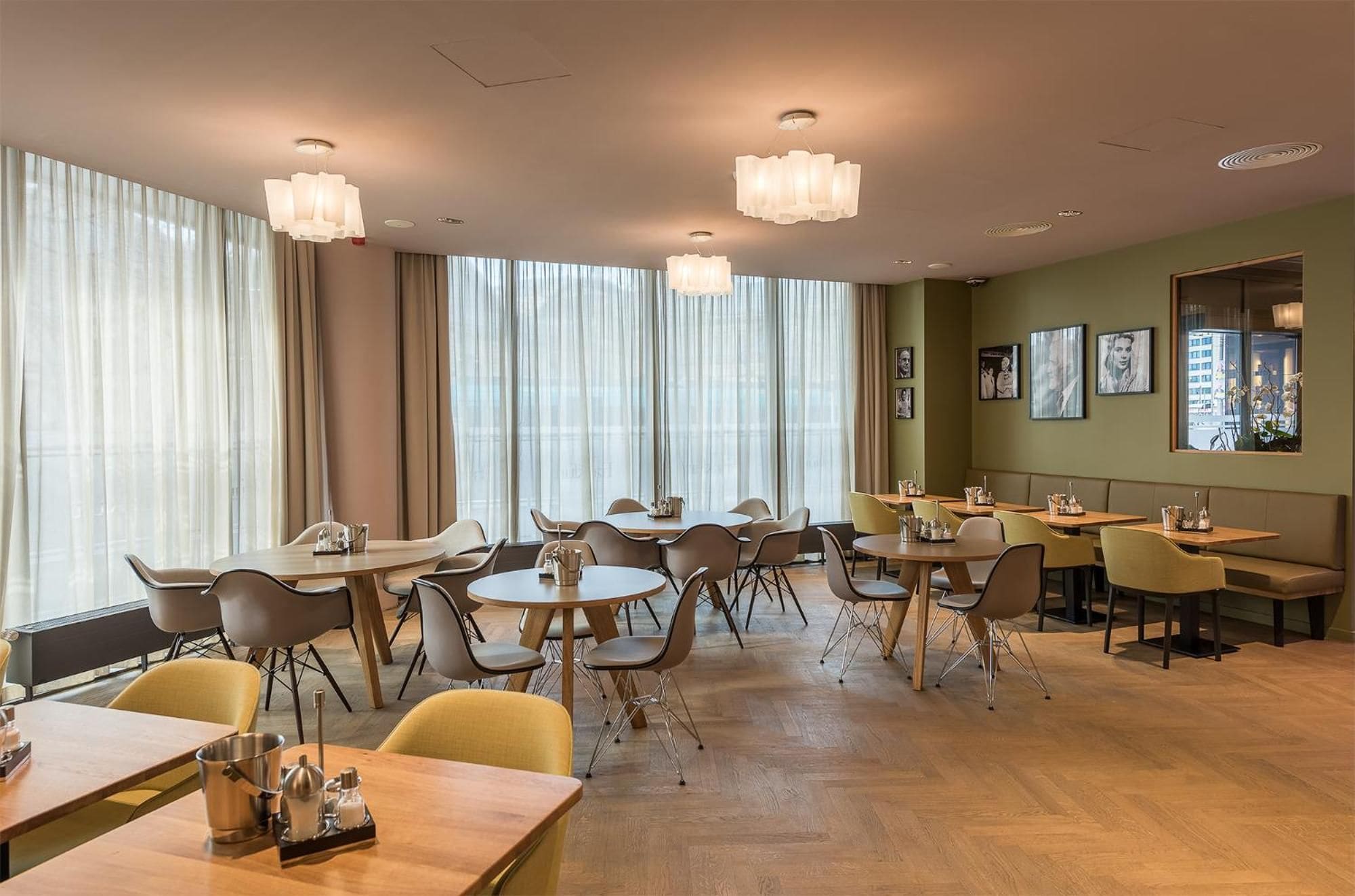 Ein Hotel-Essbereich mit mehreren Tischen und Stühlen, großen Fenstern mit Vorhängen und Bilderrahmen an der Wand.