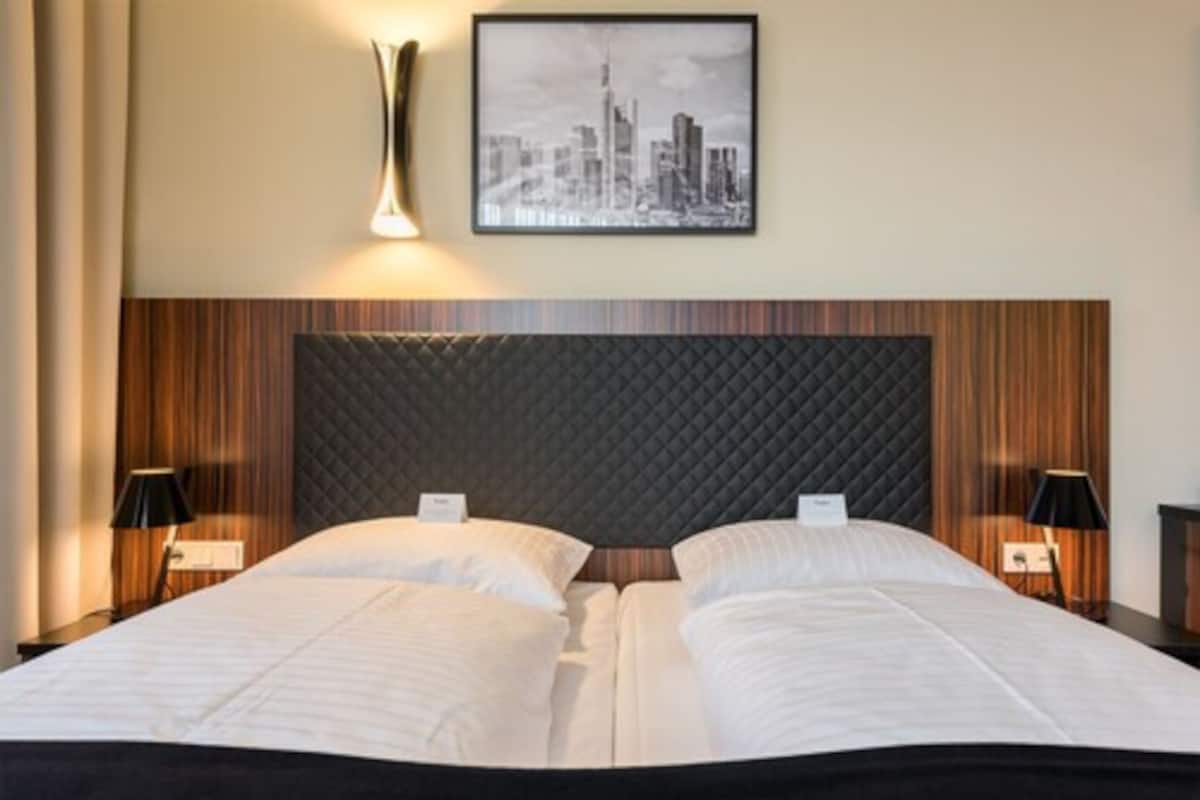 A hotel room with a large bed, two bedside tables, a cityscape artwork on the wall, and a modern headboard.