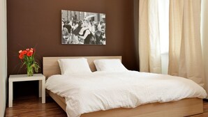 A bedroom with a wooden bed, white bedding, a small bedside table with a vase of flowers, and a black-and-white photograph on the wall. - Grampa's Hostel (Wroclaw)