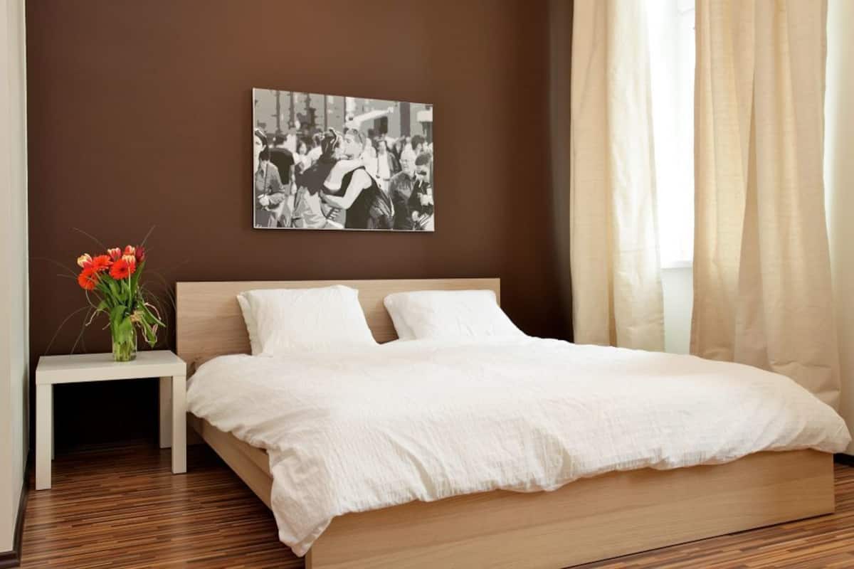 Un dormitorio con cama de madera, ropa de cama blanca, una mesita de noche con un jarrón de flores y una fotografía blanco y negro en la pared.