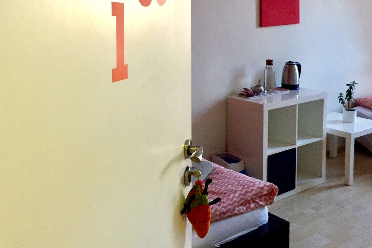 Una habitación con una repisa montada en la pared, una cama con colcha blanca y rosa estampada, una mesita con planta en maceta y un cuadro rojo en la pared.