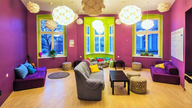 A vibrant living room with purple walls, a grey sofa, and a yellow pillow. There are two windows with green frames, a guitar, and a whiteboard on the wall. - Grampa's Hostel (Wroclaw)