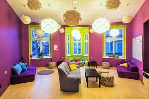 A vibrant living room with purple walls, a grey sofa, and a yellow pillow. There are two windows with green frames, a guitar, and a whiteboard on the wall. - Grampa's Hostel (Wroclaw)