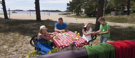 A group of people dining at an outdoor table with a red and white polka dot tablecloth, surrounded by trees and a beach in the background.