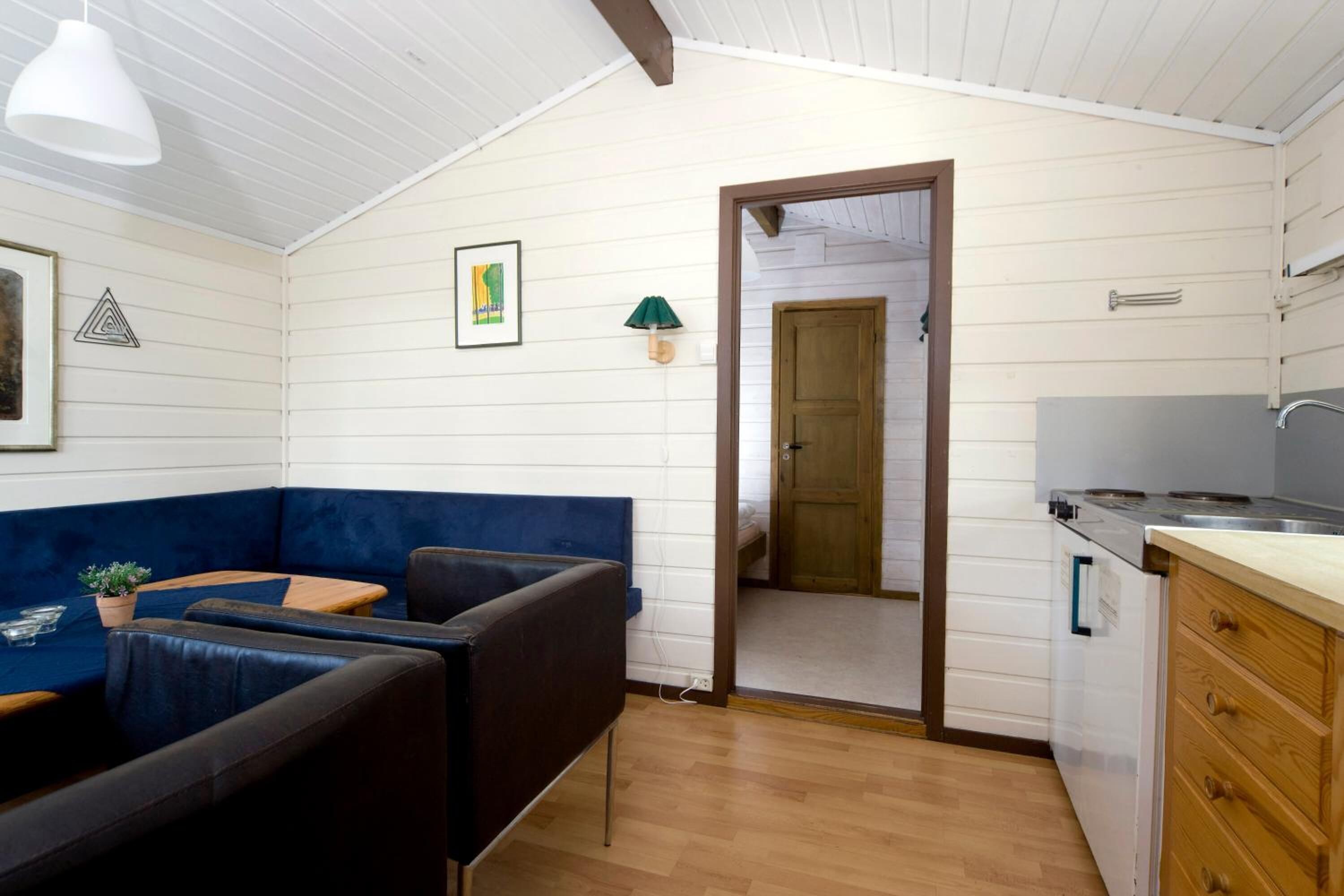 Habitació compacta amb un sofà blau, una taula de menjador de fusta, una cuineta amb fogó i una porta que condueix a una altra habitació.