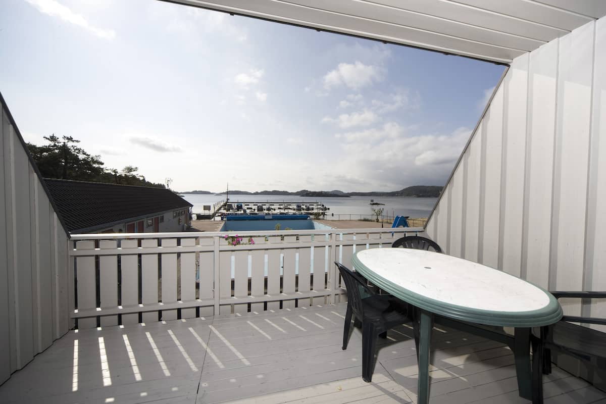 Un balcon avec une balustrade blanche, une table et des chaises. On aperçoit un bateau au loin.