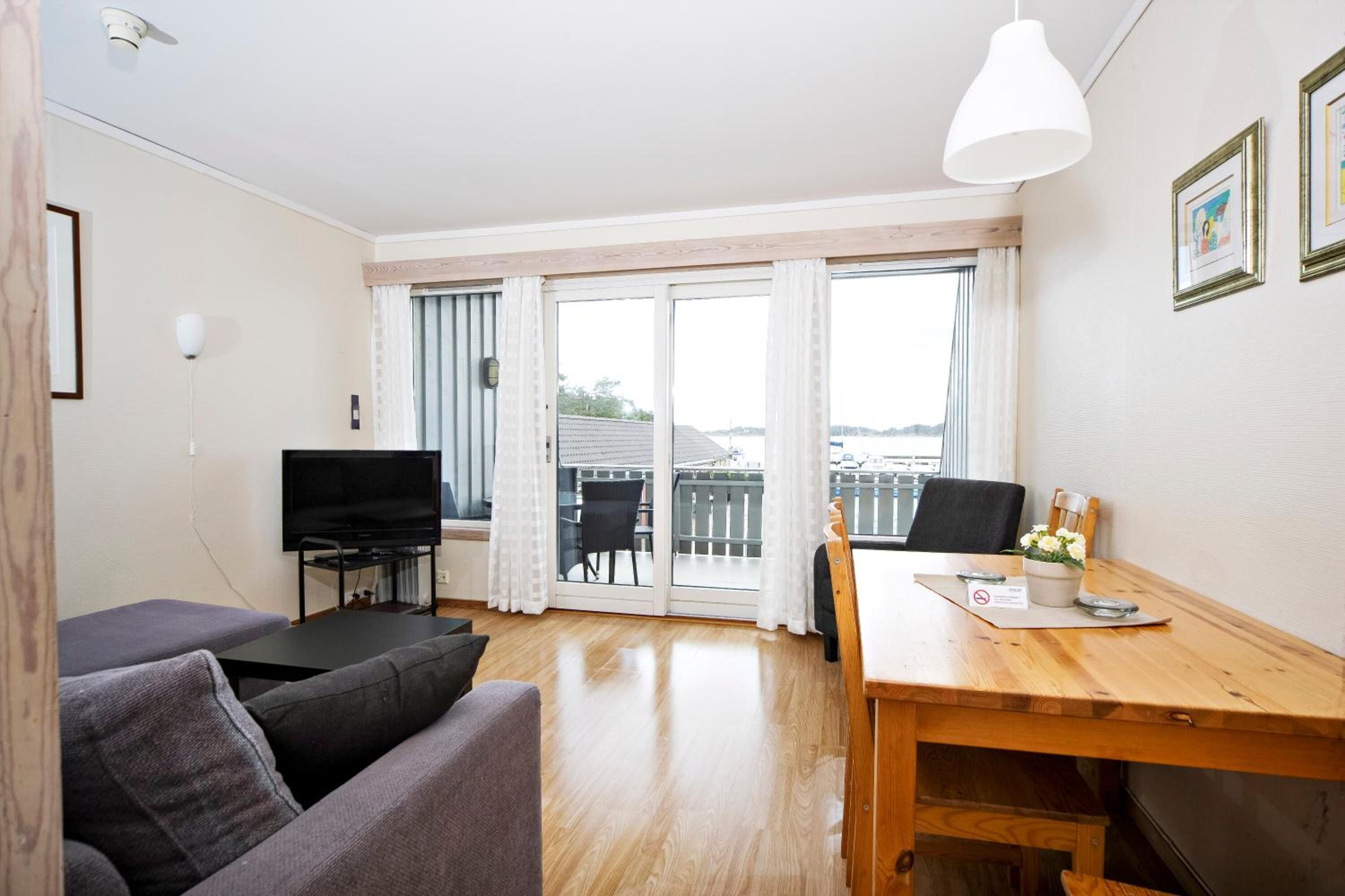 A modern living room with a sofa, a TV, a wooden dining table, and a balcony with a view.