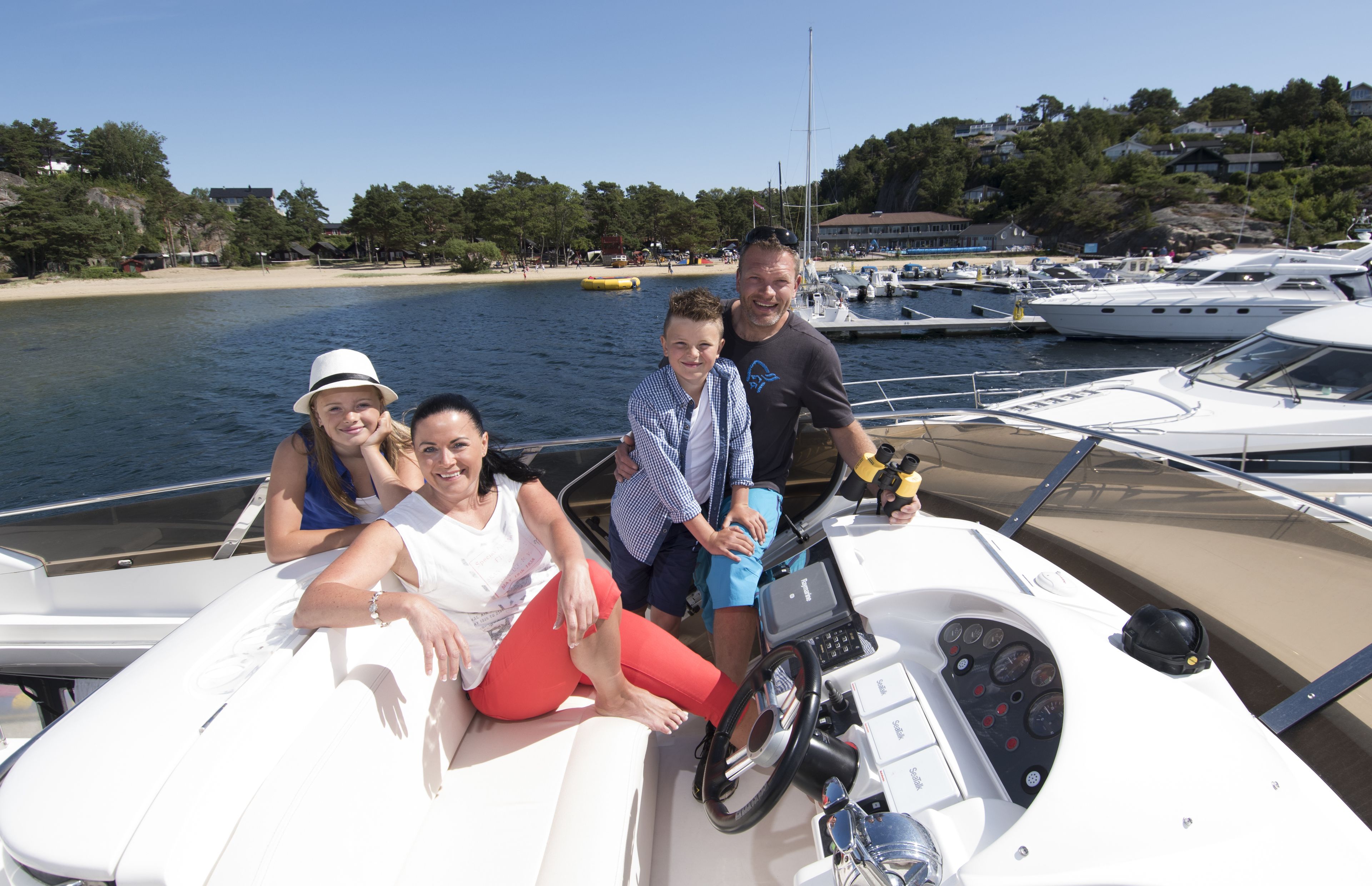 A group of people on a boat near a marina with docked yachts and a beach in the background.